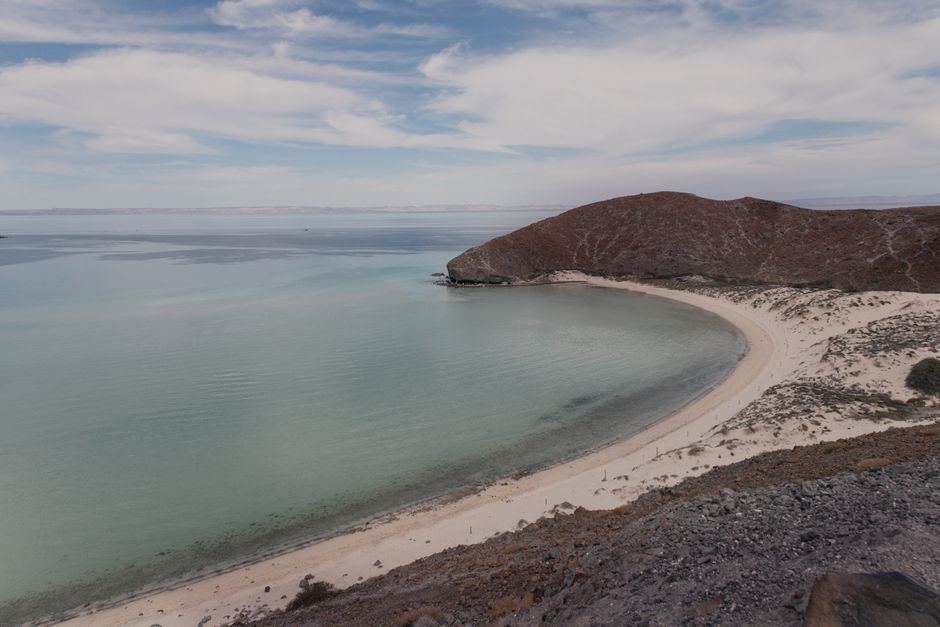 Playa de arena clara en forma de media luna junto a un mar de aguas tranquilas y un cerro rojizo, en Baja California Sur, sin personas