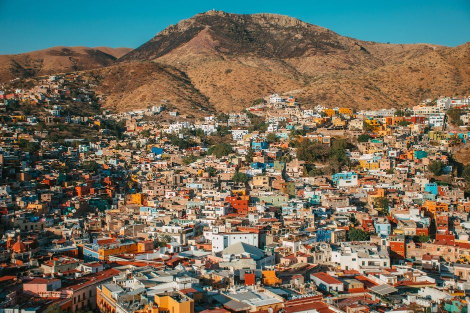 Vista aérea de casas de colores vivos sobre una ladera, con un cerro seco al fondo y cielo azul