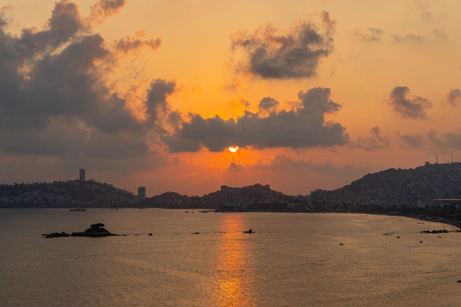 Atardecer anaranjado sobre una bahía tranquila, con nubes oscuras y colinas en siluetas al fondo