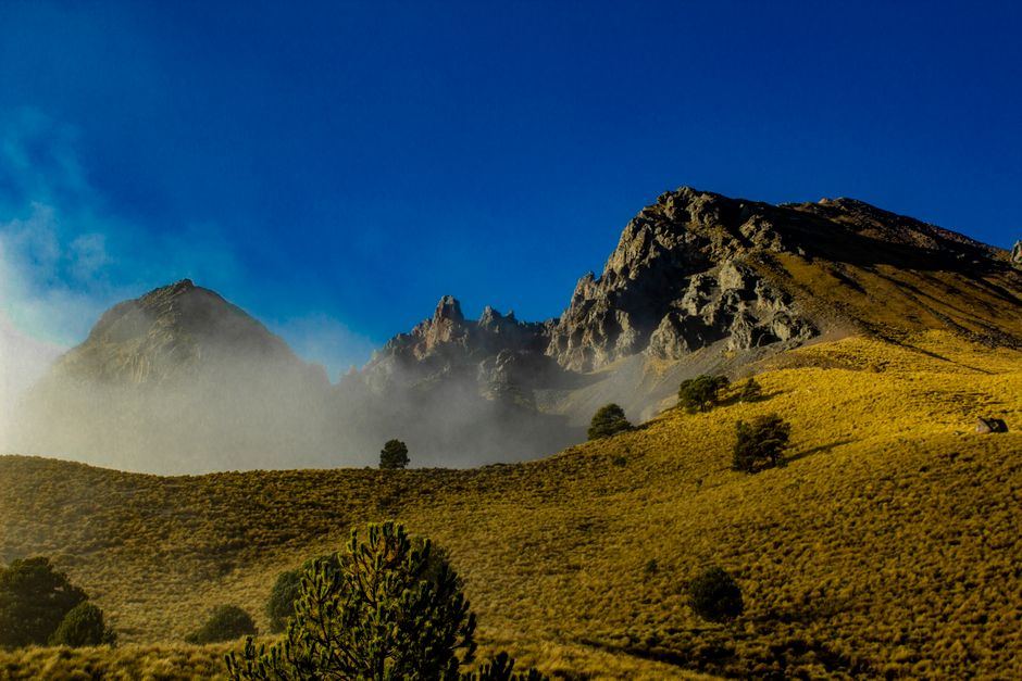 Montaña de roca y pastizal dorado con neblina bajo un cielo azul intenso, sin personas