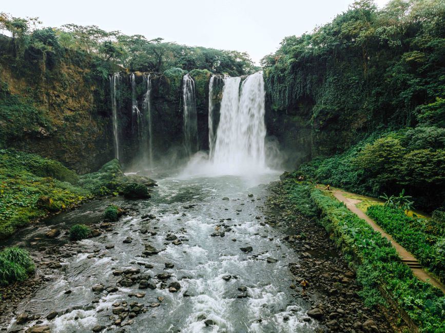Cascada ancha entre paredes cubiertas de vegetación, con un río de piedras en primer plano, sin personas