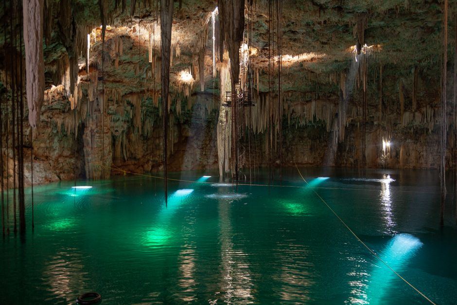 Cenote de aguas verdes iluminadas dentro de una cueva con estalactitas y haces de luz, sin personas