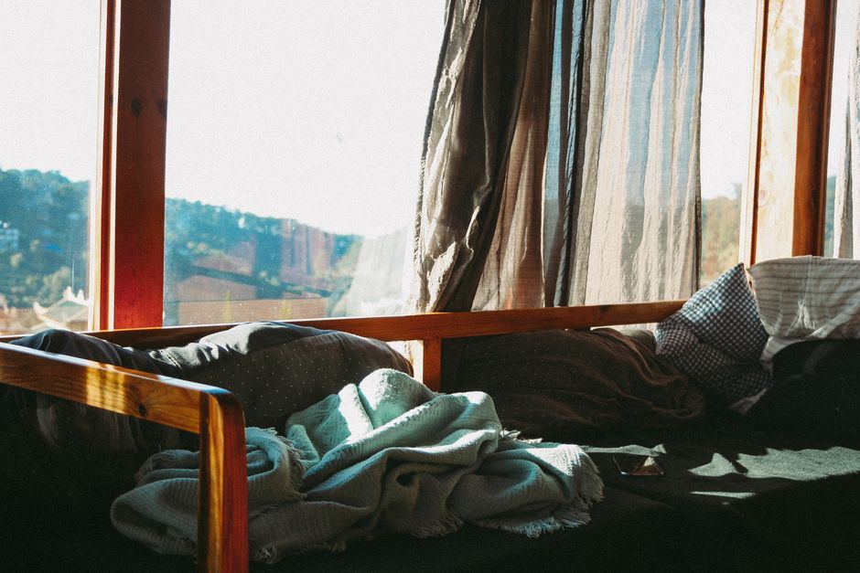 Un sillón junto a una ventana con una manta doblada y luz de la mañana entrando por una cortina