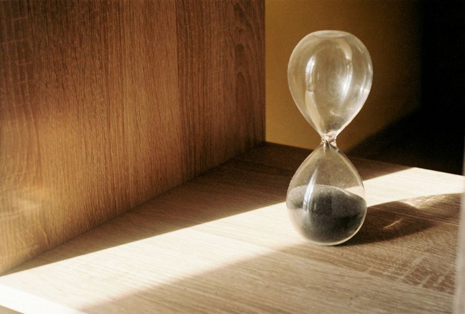 Reloj de arena con arena negra sobre una repisa de madera, bajo un rayo de luz cálida