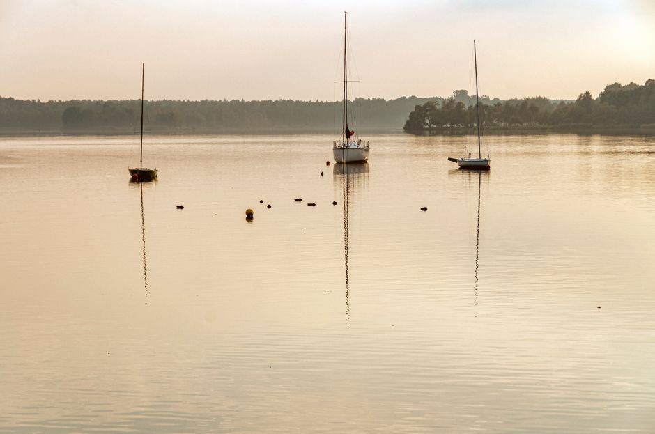 Lago en calma al amanecer con tres veleros a lo lejos y reflejos dorados en el agua, sin personas