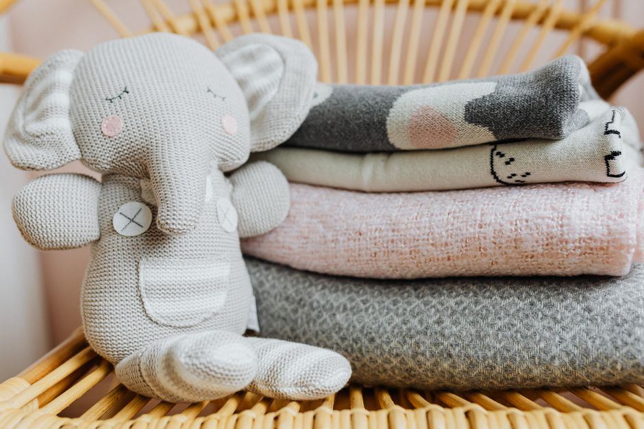 Peluche de punto gris con forma de elefante junto a mantas dobladas de colores suaves sobre una silla de mimbre, sin personas