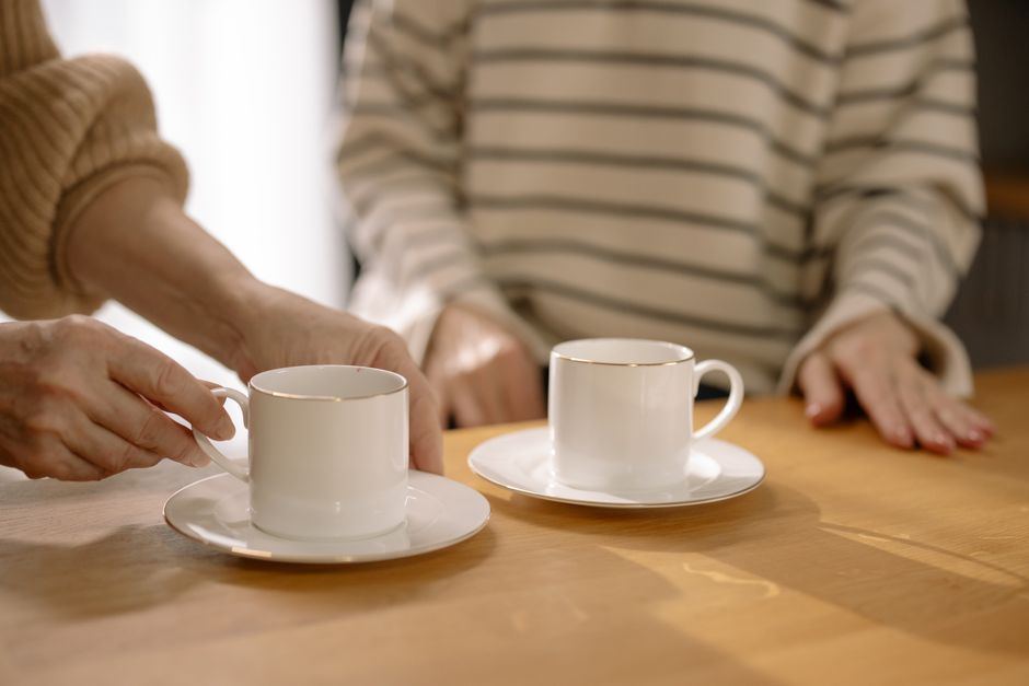 Dos tazas blancas sobre una mesa de madera clara y las manos de dos personas, sin rostros ni texto
